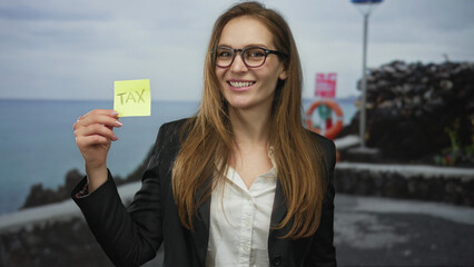 Woman wearing glasses holds yellow sticky note on seaside street by a lifebuoy and smiles broadly;...