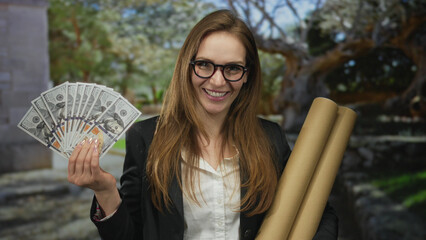 Woman holding fanned hundred dollar bills and cardboard tubes smiles in a forest setting;...