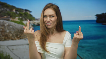 Woman extends both middle fingers toward camera at bright sandy beach with clear rocky coast;...