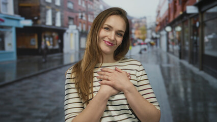 Young caucasian woman holds hands on chest standing on wet city street with blurred storefronts behind; gratitude.