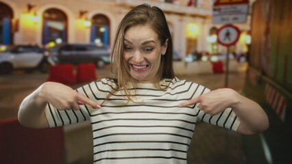 Young caucasian woman wearing striped shirt points finger to chest in illuminated urban street at...