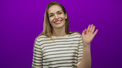 Young caucasian woman waves hand in purple studio setting with bright lighting and casual striped...