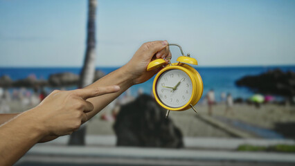 Male hand holding yellow alarm clock at beach with blurred seaside background under blue sky...