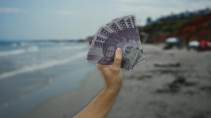 Man holding indonesian rupiah banknotes on a seaside beach setting, showcasing a concept of wealth...