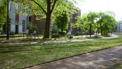 Blurred park scene with bokeh effect featuring people walking and cycling under lush green trees on a sunny day creating a peaceful urban atmosphere.