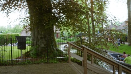 Blurred background of giethoorn canals in netherlands with defocused people relaxing by the water amidst lush greenery and a charming wooden bridge.