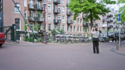 Fototapeta premium Woman photographing blurred european street with bicycles and brick buildings in an urban city under a tree on a spring day adds charm and beauty to the outdoor scene.