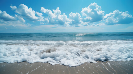 Fototapeta premium Waves crashing on the shore with a clear blue sky dotted with fluffy white clouds on a bright sunny day.