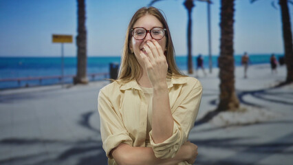 Young blonde woman covers mouth while laughing amid palm trees on a sunlit seaside promenade;...