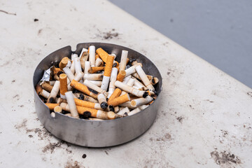 Pile of cigarette butts in a stainless steel ashtray placed located behind the building in a smoking area in outdoor with ashes and dust scattered around.
