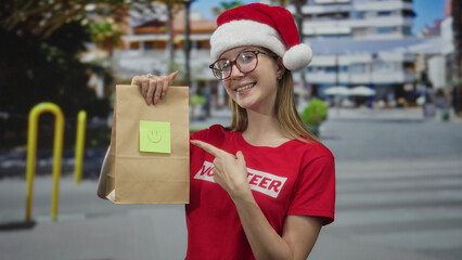 Woman wearing red volunteer shirt and santa hat finger pointing to paper bag on street; compassion...