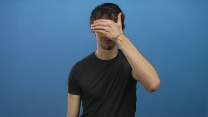Man with furrowed brow covers eyes with hand over face wearing black shirt against blue studio...