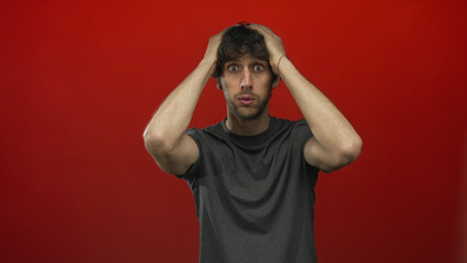 Young man holding head with both hands in bright red studio with wide eyes and tense posture;...