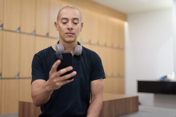Hispanic man using smartphone while seated in locker room, gym workout break, fitness lifestyle concept