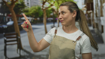Middle aged blonde woman smiling and pointing finger along a sunlit sidewalk on a city street;...