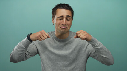 Young hispanic man wearing wristwatch holds fists to eyes in crying gesture inside blue studio set;...