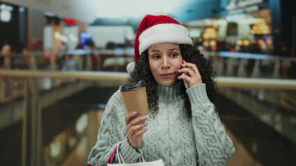 Hispanic woman in a santa hat holding coffee talks on phone while shopping indoors at a bustling...
