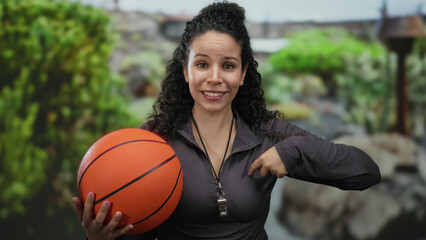 Young hispanic woman in park holding basketball and wearing whistle in casual outfit pointing at herself with trees in background outdoors