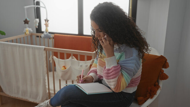 Young hispanic woman talking on phone while writing in a notebook in a cozy bedroom with a baby crib nearby, showcasing a peaceful home interior setting for multitasking mothers.