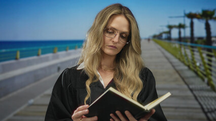 Young blonde woman in judge uniform reading a book on a seaside promenade, embodying a serene...