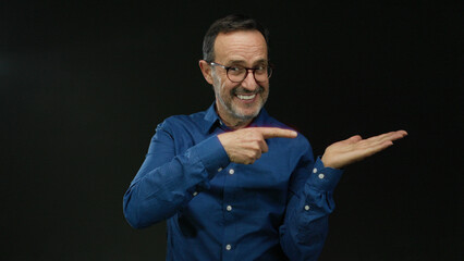 Hispanic man with glasses in blue shirt gesturing over open palm against isolated black background suggesting offering or displaying something with a confident expression