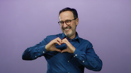 Hispanic man in blue shirt making heart gesture against purple background, expressing happiness and love.
