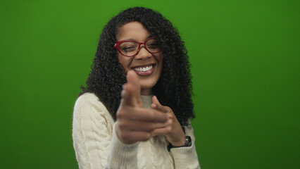 Woman with curly hair and red glasses smiling and pointing finger at camera wearing white sweater in green studio; confidence.