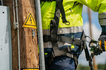 Utility worker in safety harness climbing wooden electric pole with high voltage warning sign...