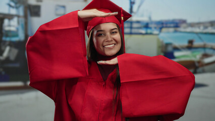 Woman in red graduation gown smiling joyfully at seaside port, showcasing achievement with boats...