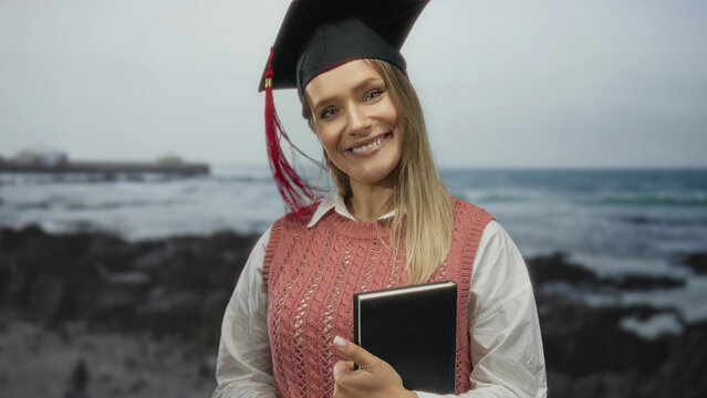 Young woman celebrating graduation at seaside holding book with blonde hair and cap smiling outdoors at the beach with ocean waves in the background