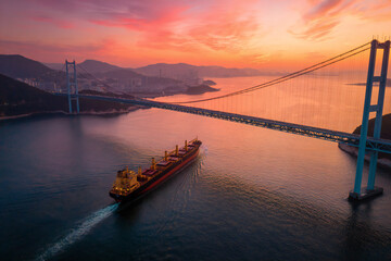 A cargo ship passing under a large suspension bridge at sunrise, calm waters, industrial and natural contrast
