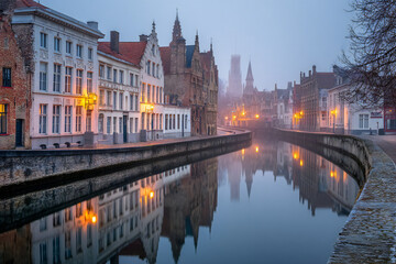 Fototapeta premium A foggy canal in the early morning in Bruges, with still water and historic buildings on both sides, moody atmosphere