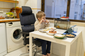 Little boy eating lunch and watching videos on tablet in kitchen
