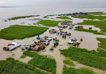 Sariakandi, Bangladesh - 15 July 2019: Aerial view of homes submerged in floodwaters amidst vibrant green vegetation, reflecting the sky's muted tones in the still, encompassing water..