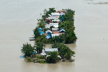 Sariakandi, Bangladesh - 15 July 2019: Aerial view of a village, homes dotted with red and blue roofs, barely rising above the floodwaters, reflecting the sky's muted tones.