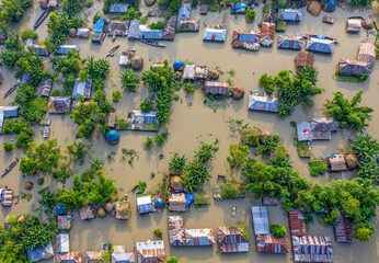 Sariakandi, Bangladesh - 15 July 2019: Aerial view of houses submerged in floodwater, where roofs peek through the murky water, interspersed with verdant trees, under a hazy sky.