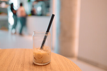 Partially finished glass of iced coffee with a black straw on a wooden table inside a bright modern cafe. The background shows people near the counter out of focus, creating a casual and relaxed