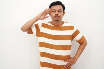 Young Asian man in white and brown striped t-shirt stands saluting with a serious expression,...