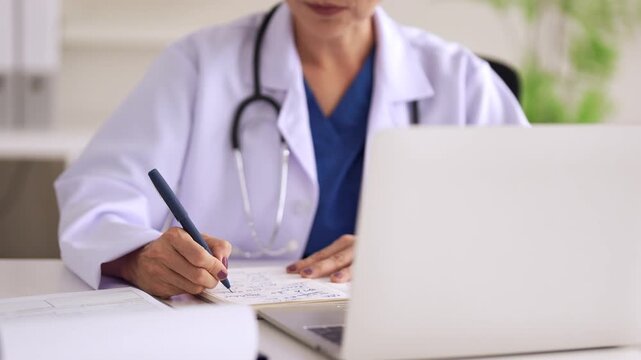 Asian adult female doctor writing medical notes on a chart in a clinic office for patient healthcare administration and professional record keeping