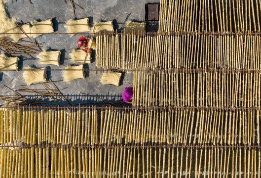Bogura, Bangladesh - 22 April 2021: Aerial view of golden noodles drying in precise rows under the sun, contrasting with the vibrant magenta and red of the workers' attire.