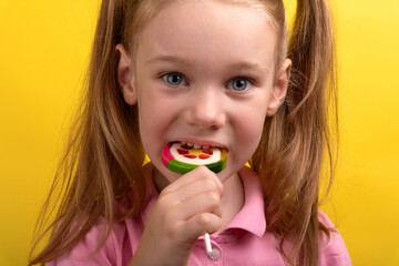 Close-up of a young girl with pigtails biting a vibrant lollipop, wearing a pink shirt against a...