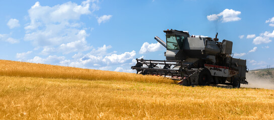 Agricultural combine harvesting golden wheat on a sloped field under a sunny sky with scattered...