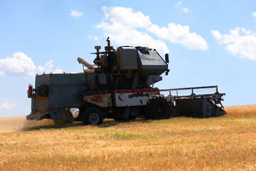 Fototapeta premium A weathered combine harvester working on a dry wheat field under a sunny blue sky with scattered clouds.