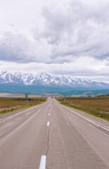 Naklejka premium Endless road in Altai region leads to towering snowy peaks. Open landscape and stormy clouds create a powerful sense of freedom and exploration in southern Siberia.