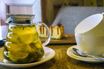 Glass teapot with green tea, mint and lemon, cup of tea, small teaspoon and half-eaten piece of cheesecake on table in cafe, food and drink. Close-up.