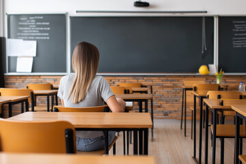 Student sitting alone at wooden desk in classroom with chalkboard and projector