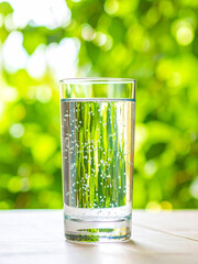 Glass of Sparkling Mineral Water with Condensation on Light Surface and Green Natural Bokeh Background