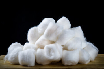 A pile of white cotton balls arranged on a wooden surface with black background. The soft texture and contrast evoke purity, hygiene, and minimalist calm.
