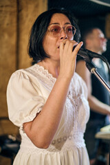 Fototapeta premium Young woman with dark hair and glasses passionately sings into microphone. Casual studio surrounds, warm lighting enhances focus on expressive performance. Background features blurred male musician