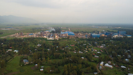 Aerial shot captures the contrast between industry and village, highlighting environmental impact near lush landscape.
